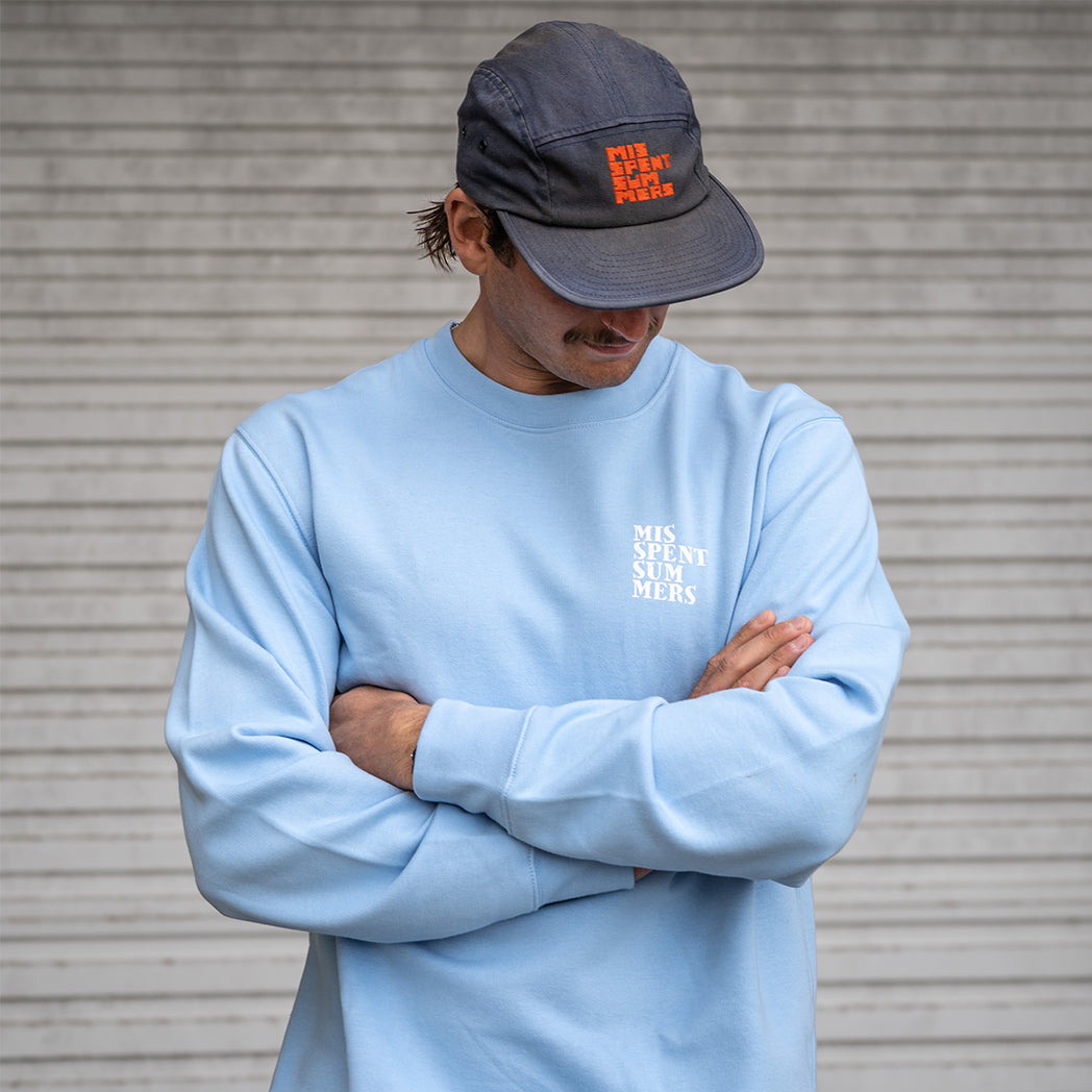 Person wearing a light blue sweatshirt and dark cap with text, standing against a metallic background.